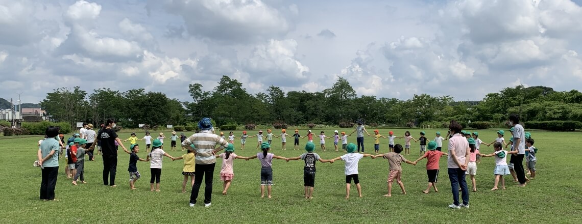 写真：芝生の広がる大きな広場にて、先生と子どもたちが手をつなぎ合って大きな円を形作っている様子