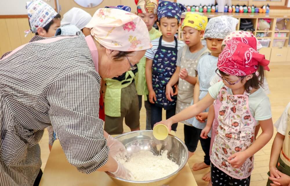 写真：料理教室で先生が手作りするのを園児たちが手伝う様子