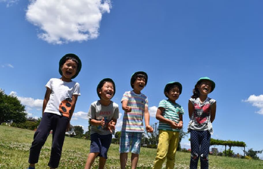 写真：青空の元、芝生が広がる広場にて、子どもたちが一列に横に並んで思いっきり笑っている様子
